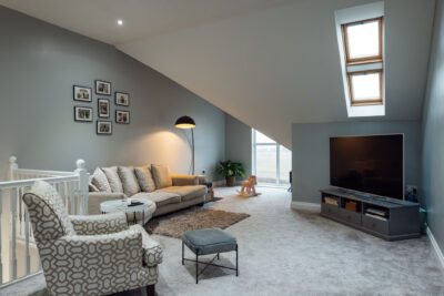 A wide view of a luxury modern living room area with a sofa, armchair and TV in the room and skylight windows - The Attic Guys